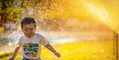 Young child running through water spray that is illuminated yellow by the sun. He is wearing a t-shirt with various positive words and he is laughing.