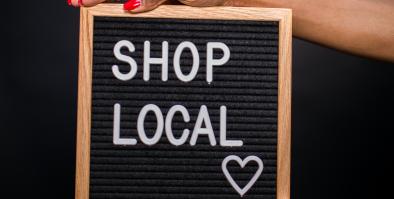 hands holding a square sign with a black background white text shop local with a heart