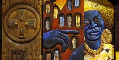 mixed media art on a carved wood base with embedded glass, metal, and painted elements showing a blue-black toned hand pointing right in the center and an image of a blue-black skinned femme at left wearing golden jewelry