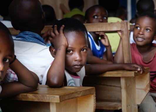 Children sitting at desks with focus on one child in the middle.  He is turned away from his desk and has his elbow on the desk of the child behind him with his head on his hand.  He is looking at the camera with a glum expression.