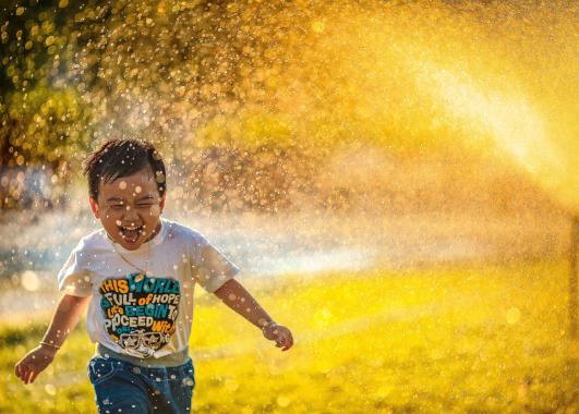 Young child running through water spray that is illuminated yellow by the sun. He is wearing a t-shirt with various positive words and he is laughing.