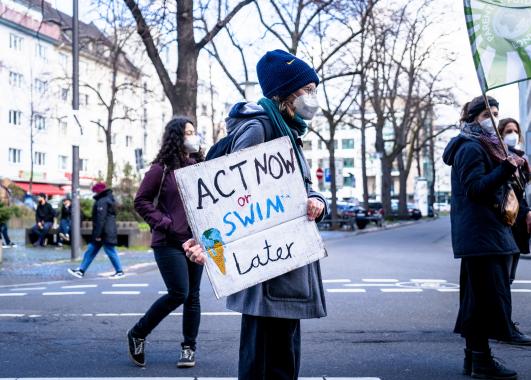 Photo of protester holding sign reading "Act Now or Swim Later"